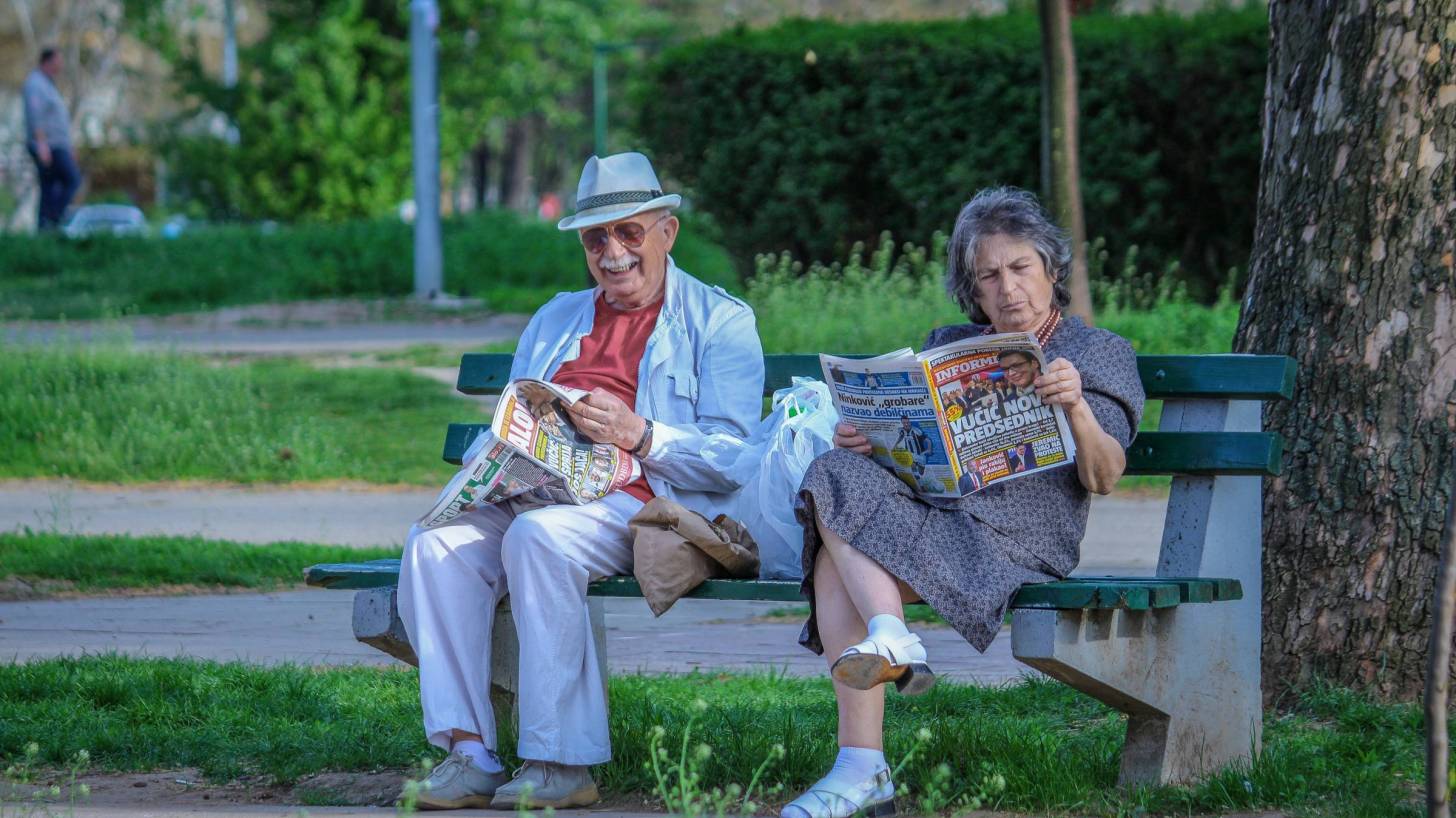 senior couple sitting on park bench reading paper